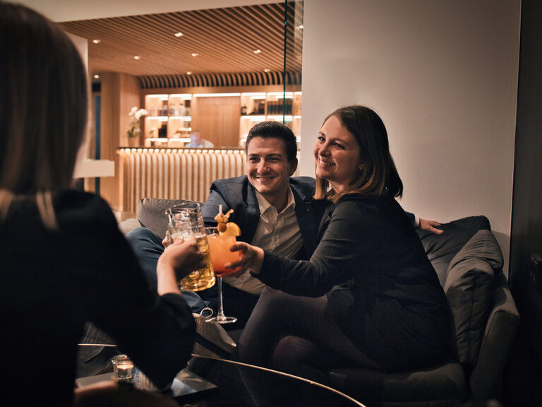 Drei Personen stoßen mit einem Drink an und genießen den Abend im Hotelbarbereich